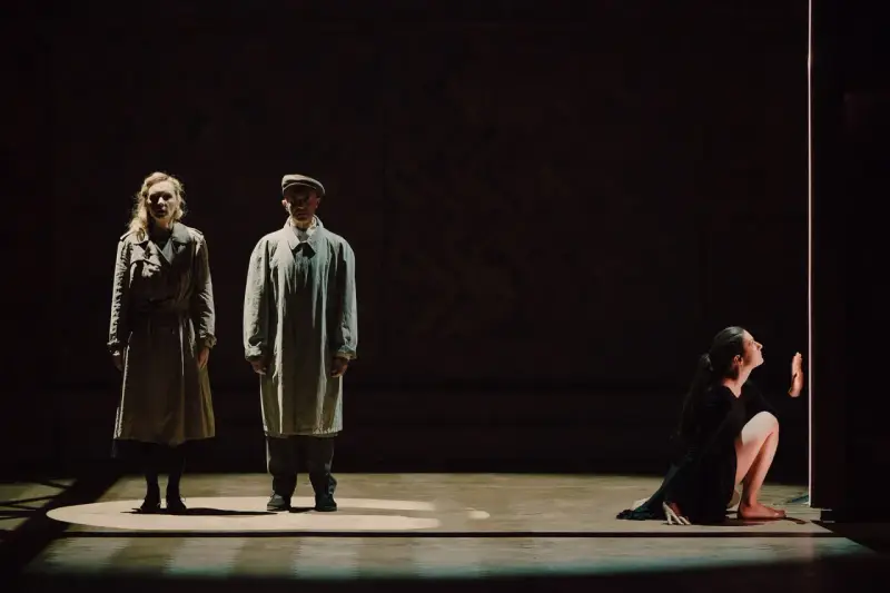 Magdalena Twardowska w scenie teatralnej. Dwie postacie w płaszczach stoją w świetle, obok nich kobieta w czarnej sukni klęczy przy pionowym świetle.